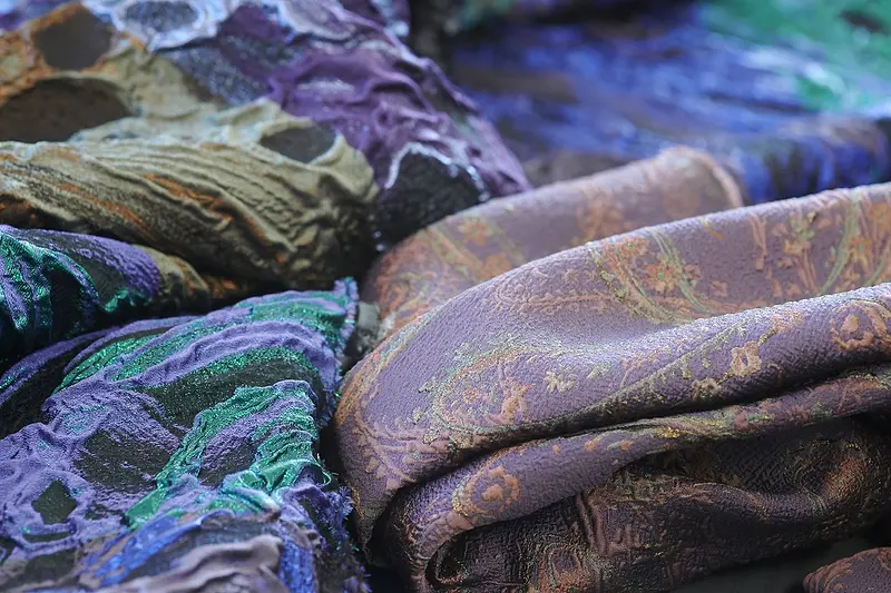 Tessuti jacquard in viola, verde e oro, con riflessi cangianti, ammucchiati sul tavolo